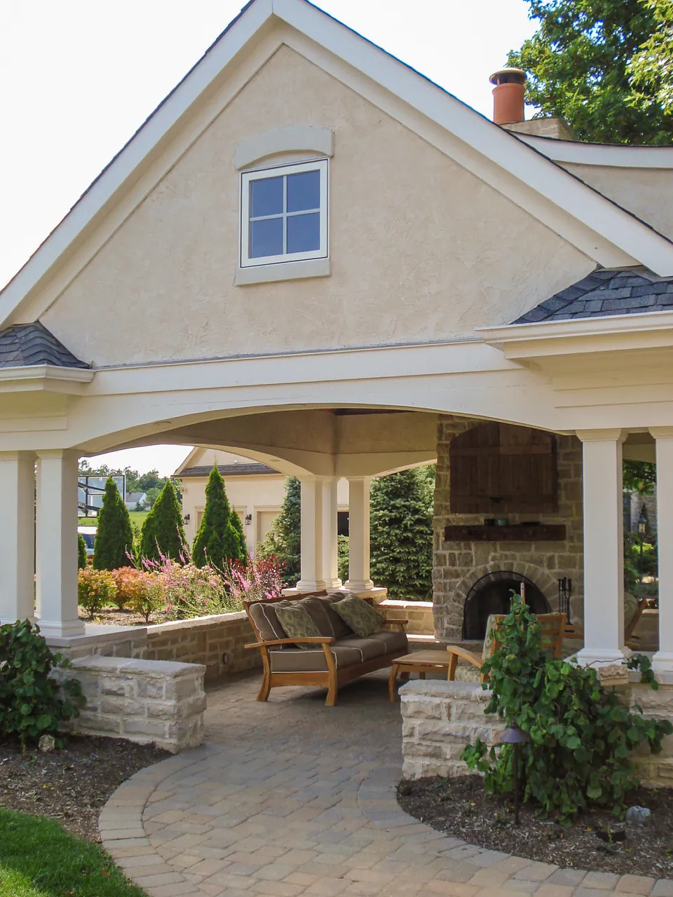 Paver walk leading toward covered pavilion with stone fireplace, columns, shrubs, and foundation planting.